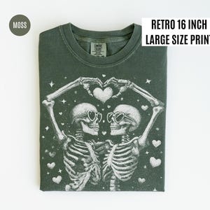 May include: Moss green t-shirt with a retro 16-inch large-size print. The graphic features two skeletons wearing heart-shaped sunglasses, forming a heart with their hands. White hearts and stars are scattered around the skeletons.