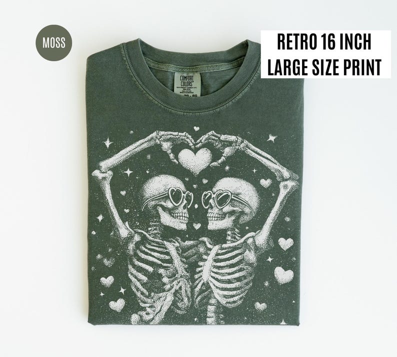 May include: Moss green t-shirt with a retro 16-inch large-size print. The graphic features two skeletons wearing heart-shaped sunglasses, forming a heart with their hands. White hearts and stars are scattered around the skeletons.