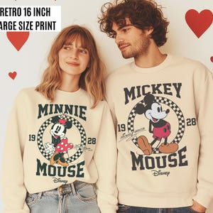 Może przedstawiać: Kremowe bluzy z nadrukami Myszki Miki i Myszki Minnie. Bluzy mają czarny tekst i grafikę, w tym słowa "Mickey Mouse" i "Minnie Mouse". U g&oacute;ry znajduje się tekst "Retro 40.6 cm duży nadruk".