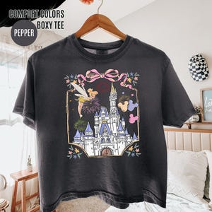 May include: A dark gray boxy tee shirt with a colorful graphic of a castle, fireworks, and Tinkerbell. The graphic is framed with a pink bow and floral accents. The shirt has the text "COMFORT COLORS BOXY TEE" and "PEPPER".