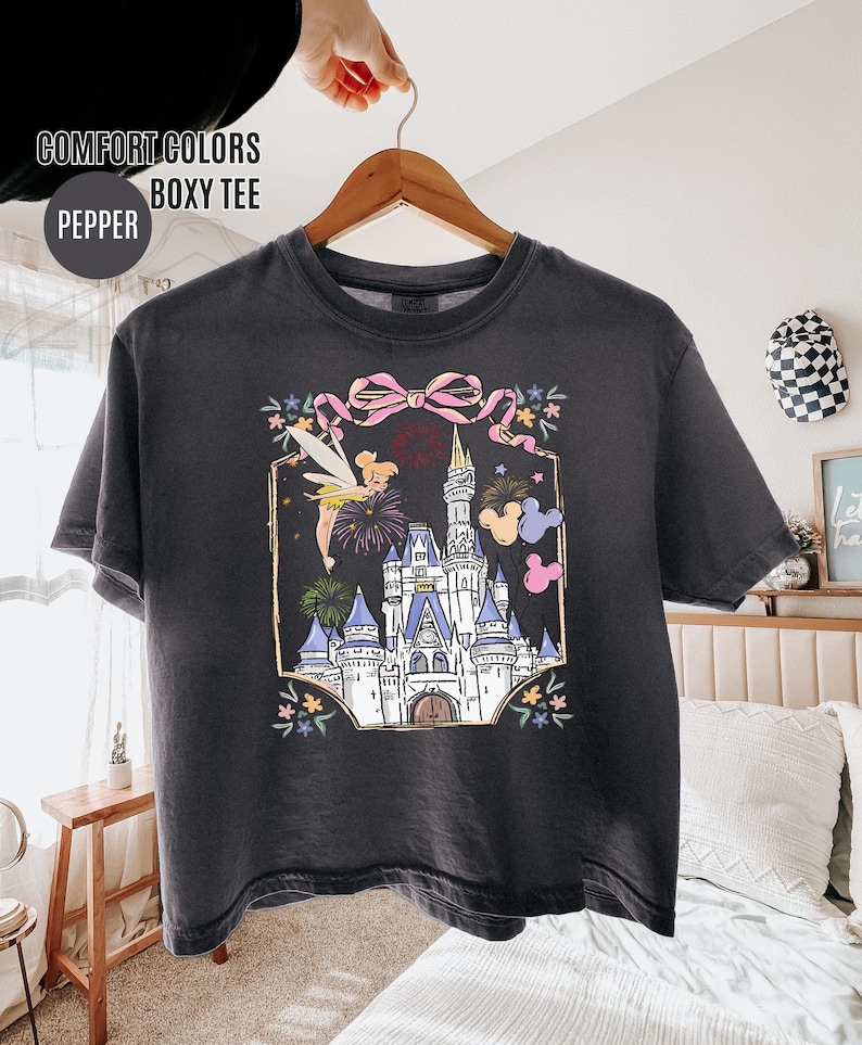 May include: A dark gray boxy tee shirt with a colorful graphic of a castle, fireworks, and Tinkerbell. The graphic is framed with a pink bow and floral accents. The shirt has the text "COMFORT COLORS BOXY TEE" and "PEPPER".