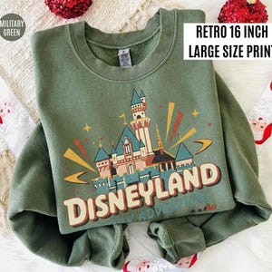 May include: Military green sweatshirt with a retro Disneyland castle graphic. The design includes the text "Disneyland" in a vintage font, with a large size print.