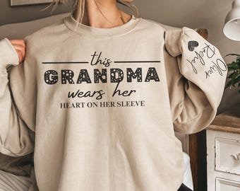 May include: A beige sweatshirt with the text "this grandma wears her heart on her sleeve" in black. The text is decorated with small black dots. The sleeve has a black heart with the text "Grandma" written inside.