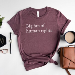 May include: A maroon t-shirt with the text "Big fan of human rights." is displayed on a white surface. The shirt is styled with a knot at the bottom. Accessories include a watch, glasses, earrings, a cup of coffee, and a brown purse.