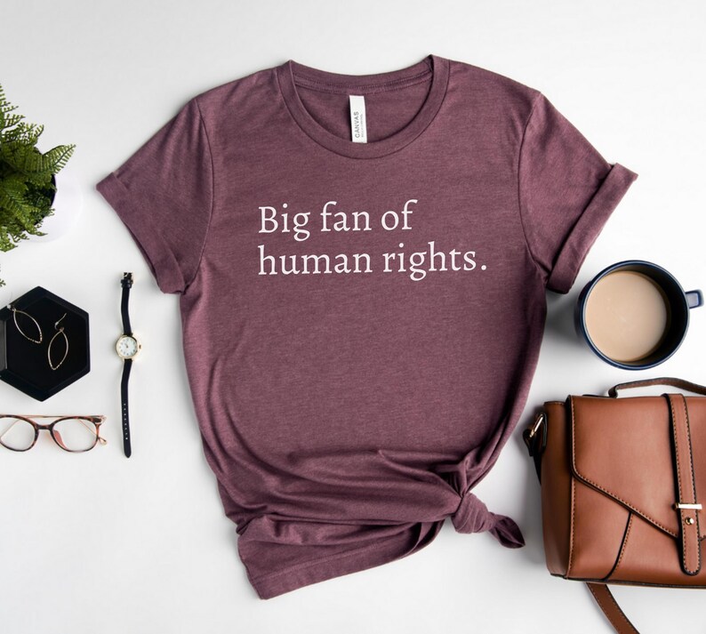 May include: A maroon t-shirt with the text "Big fan of human rights." is displayed on a white surface. The shirt is styled with a knot at the bottom. Accessories include a watch, glasses, earrings, a cup of coffee, and a brown purse.