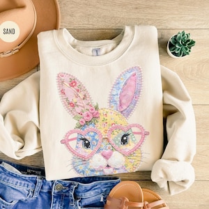 Può includere: Una felpa color crema con un volto di coniglio colorato con occhiali a forma di cuore. Il design del coniglio incorpora motivi floreali in rosa, blu e giallo. La parola "SAND" è stampata su un cappello marrone nell'angolo in alto a sinistra.