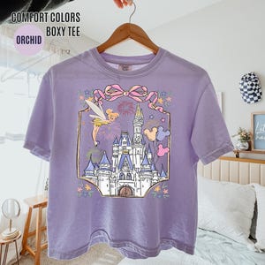 May include: Orchid-colored boxy tee shirt featuring a graphic of a fairytale castle, fireworks, and a fairy. The design includes a pink bow and Mickey Mouse balloons. The shirt is hanging on a wooden hanger.