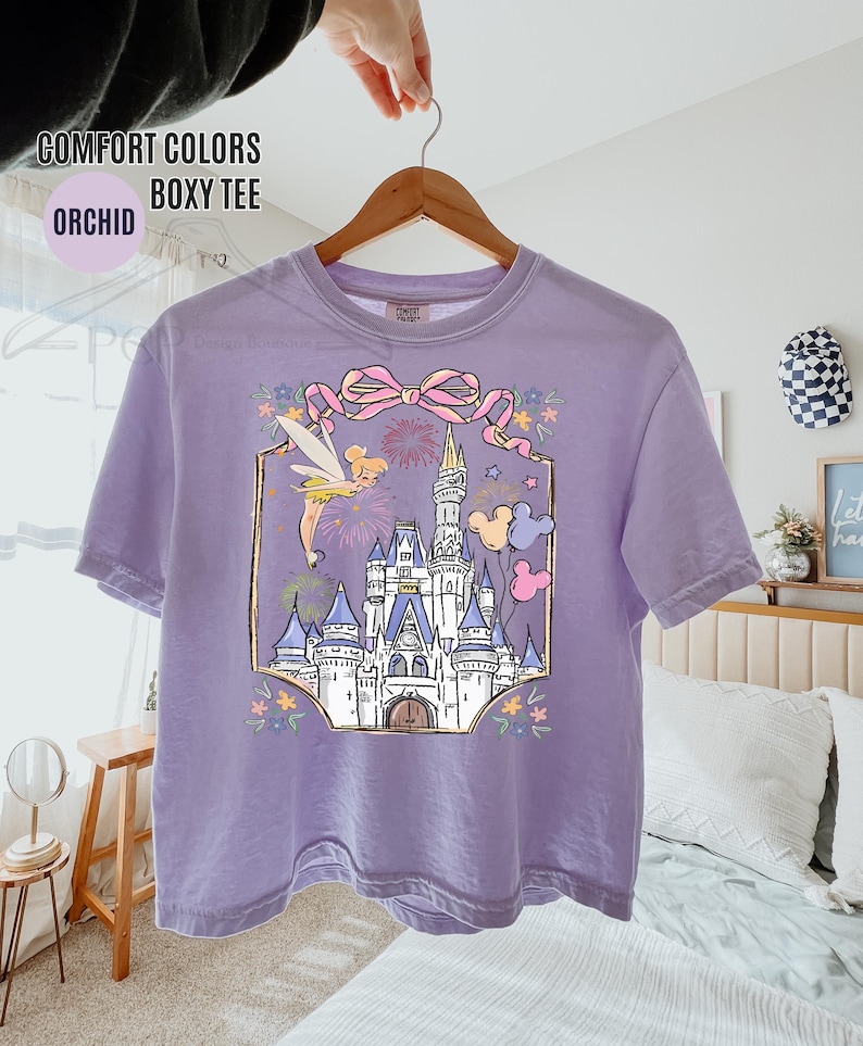 May include: Orchid-colored boxy tee shirt featuring a graphic of a fairytale castle, fireworks, and a fairy. The design includes a pink bow and Mickey Mouse balloons. The shirt is hanging on a wooden hanger.