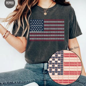 Peut inclure: T-shirt gris foncé avec un motif de drapeau américain. Le drapeau est composé du texte "FDT" dans le champ bleu et des mots "Freedom Democracy Truth" dans les rayures rouges. Le t-shirt a des manches courtes et un col rond.