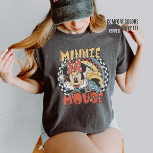 Może przedstawiać: Ciemnoszara koszulka z kolorową grafiką Myszki Minnie. Projekt zawiera twarz Minnie, czerwoną kokardę w kropki oraz napis "MINNIE MOUSE" w stylu retro. Koszulka ma pudełkowy kr&oacute;j.