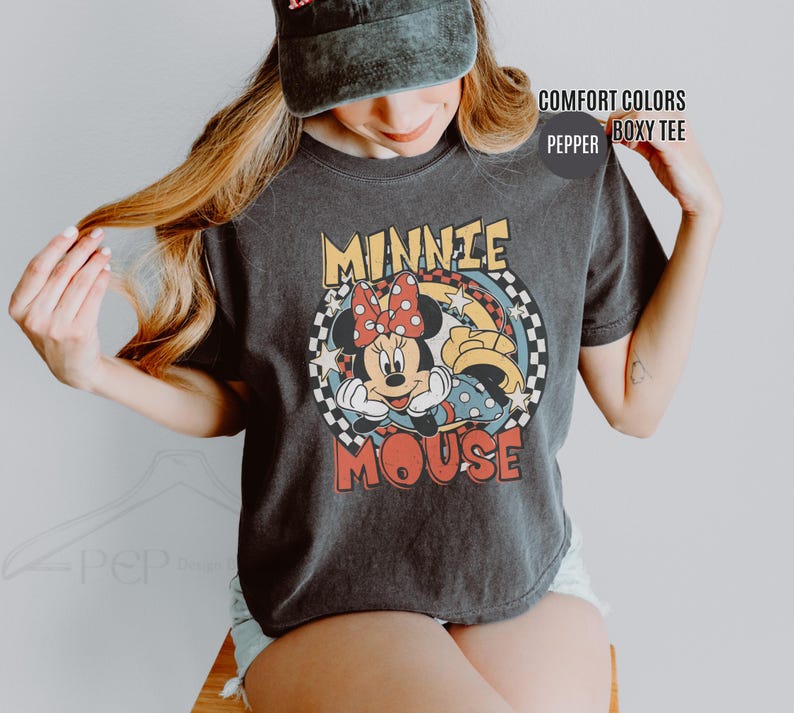 Może przedstawiać: Ciemnoszara koszulka z kolorową grafiką Myszki Minnie. Projekt zawiera twarz Minnie, czerwoną kokardę w kropki oraz napis "MINNIE MOUSE" w stylu retro. Koszulka ma pudełkowy kr&oacute;j.