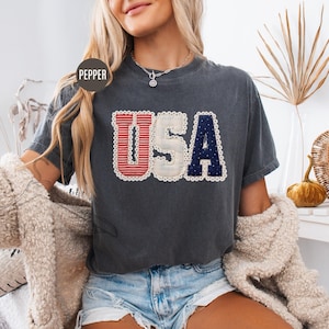 May include: A charcoal grey short-sleeve t-shirt featuring the letters USA in a large, decorative font. The U is red with white horizontal stripes, the S is off-white, and the A is navy blue with white dots. Each letter has a white lace trim.