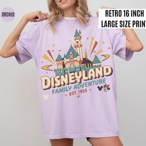 Puede incluir: Camiseta color orqu&iacute;dea con un dise&ntilde;o gr&aacute;fico retro de Disneyland. El dise&ntilde;o presenta un castillo, las palabras "DISNEYLAND FAMILY ADVENTURE + EST. 1955 +" y Mickey y Minnie Mouse. La camiseta tiene una etiqueta "RETRO 16 INCH LARGE SIZE PRINT".