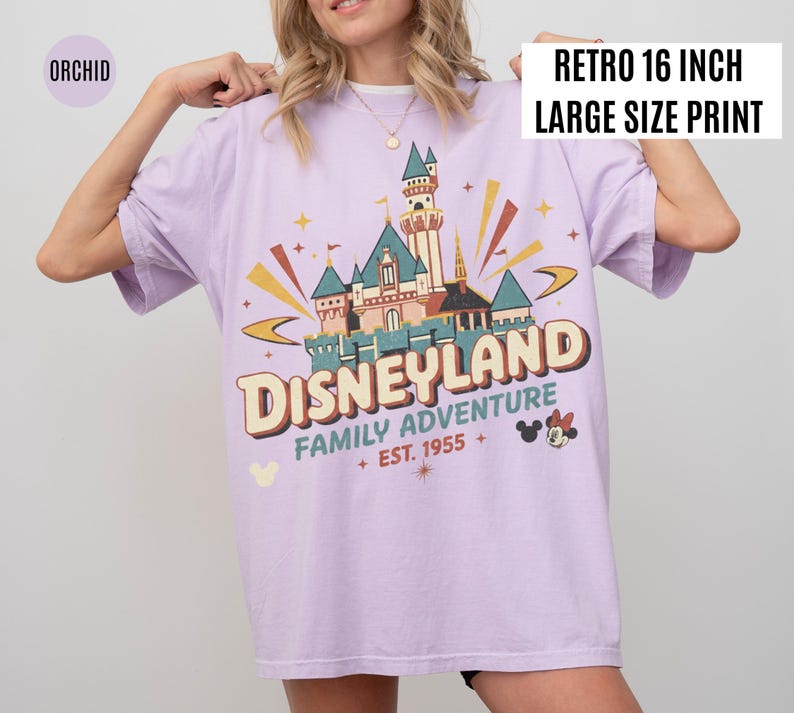 Puede incluir: Camiseta color orqu&iacute;dea con un dise&ntilde;o gr&aacute;fico retro de Disneyland. El dise&ntilde;o presenta un castillo, las palabras "DISNEYLAND FAMILY ADVENTURE + EST. 1955 +" y Mickey y Minnie Mouse. La camiseta tiene una etiqueta "RETRO 16 INCH LARGE SIZE PRINT".