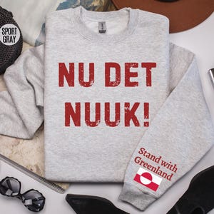 May include: A light gray sweatshirt with the text "NU DET NUUK!" in red, distressed lettering. The sleeve features the text "Stand with Greenland" and a Greenlandic flag. The sweatshirt is a sport gray color.