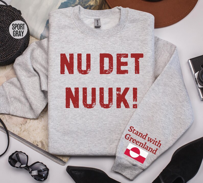 May include: A light gray sweatshirt with the text "NU DET NUUK!" in red, distressed lettering. The sleeve features the text "Stand with Greenland" and a Greenlandic flag. The sweatshirt is a sport gray color.