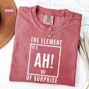May include: Crimson t-shirt with the text "THE ELEMENT AH! OF SURPRISE" in a white square. The shirt has a crew neck and a small tag. A silver necklace with a pendant is on the shirt.