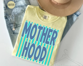 May include: A pale yellow t-shirt featuring the word "MOTHERHOOD" in blue block letters, set against a background of vertical teal stripes. The shirt is folded on top of a pair of blue jeans and a tan hat.