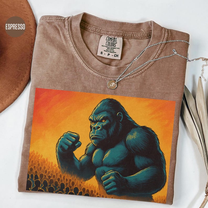 May include: A brown t-shirt with a graphic of a large gorilla with its fists clenched, standing in front of a crowd of people. The background is a sunset with orange and yellow hues. The t-shirt has the text "COMFORT COLORS" on the tag.