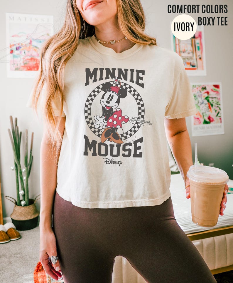 May include: Ivory-colored boxy tee featuring a graphic of Minnie Mouse in a black and white checkered circle. The text "MINNIE MOUSE" is above and below the image. The shirt also has the text "Disney" below the image.