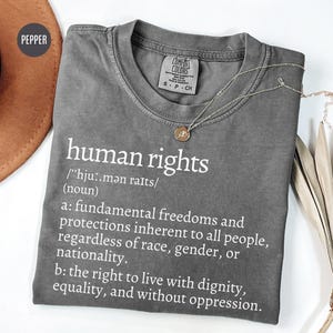 Pode incluir: Uma camiseta cinza Comfort Colors com as palavras "human rights" impressas em branco. A definição de direitos humanos também está impressa na camiseta. Um colar de prata com um pingente está na camiseta.