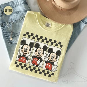 Puede incluir: Una camiseta amarilla clara con tres im&aacute;genes de Mickey Mouse en diferentes poses, enmarcadas por un patr&oacute;n de cuadros blancos y negros. La camiseta est&aacute; sobre unos vaqueros azules y un sombrero beige.