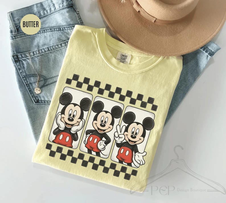 Puede incluir: Una camiseta amarilla clara con tres im&aacute;genes de Mickey Mouse en diferentes poses, enmarcadas por un patr&oacute;n de cuadros blancos y negros. La camiseta est&aacute; sobre unos vaqueros azules y un sombrero beige.