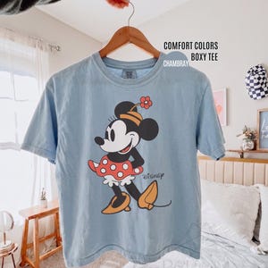 Op de afbeelding: Lichtblauw T-shirt met een afbeelding van Minnie Mouse in een rode polkadot rok en gele schoenen. Op het shirt staat de tekst "COMFORT COLORS BOXY TEE CHAMBRAY". Het T-shirt hangt aan een houten hanger.