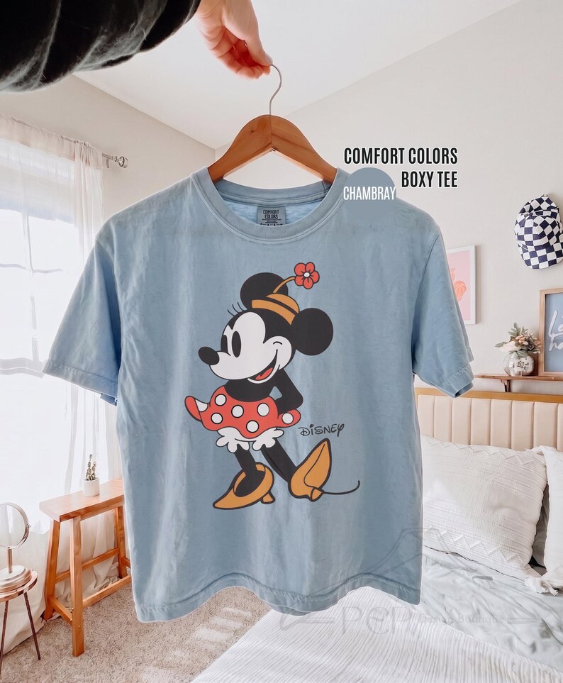 Op de afbeelding: Lichtblauw T-shirt met een afbeelding van Minnie Mouse in een rode polkadot rok en gele schoenen. Op het shirt staat de tekst "COMFORT COLORS BOXY TEE CHAMBRAY". Het T-shirt hangt aan een houten hanger.