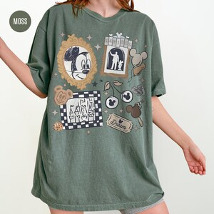 May include: Moss green t-shirt with various cartoon graphics. The shirt features a sketch of Mickey Mouse, a hotel scene, Mickey Mouse-shaped balloons, cherries, and a ticket that says "Dream".