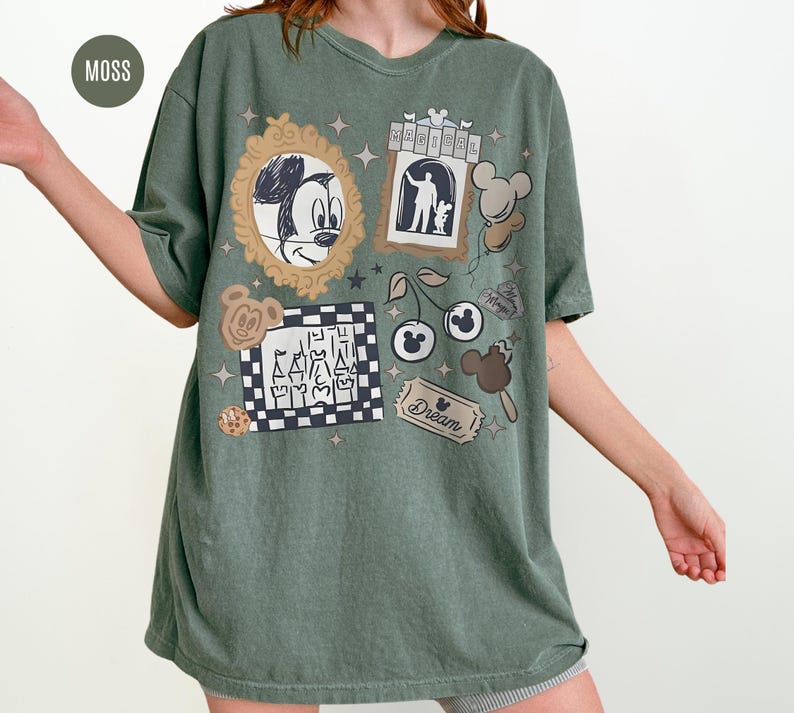 May include: Moss green t-shirt with various cartoon graphics. The shirt features a sketch of Mickey Mouse, a hotel scene, Mickey Mouse-shaped balloons, cherries, and a ticket that says "Dream".