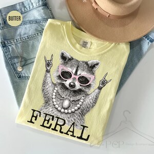 May include: A light yellow t-shirt featuring a raccoon wearing pink sunglasses and a pearl necklace, with the word "FERAL" in black block letters. The shirt is on top of blue jeans and a tan hat.