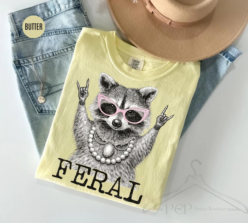 May include: A light yellow t-shirt featuring a raccoon wearing pink sunglasses and a pearl necklace, with the word "FERAL" in black block letters. The shirt is on top of blue jeans and a tan hat.