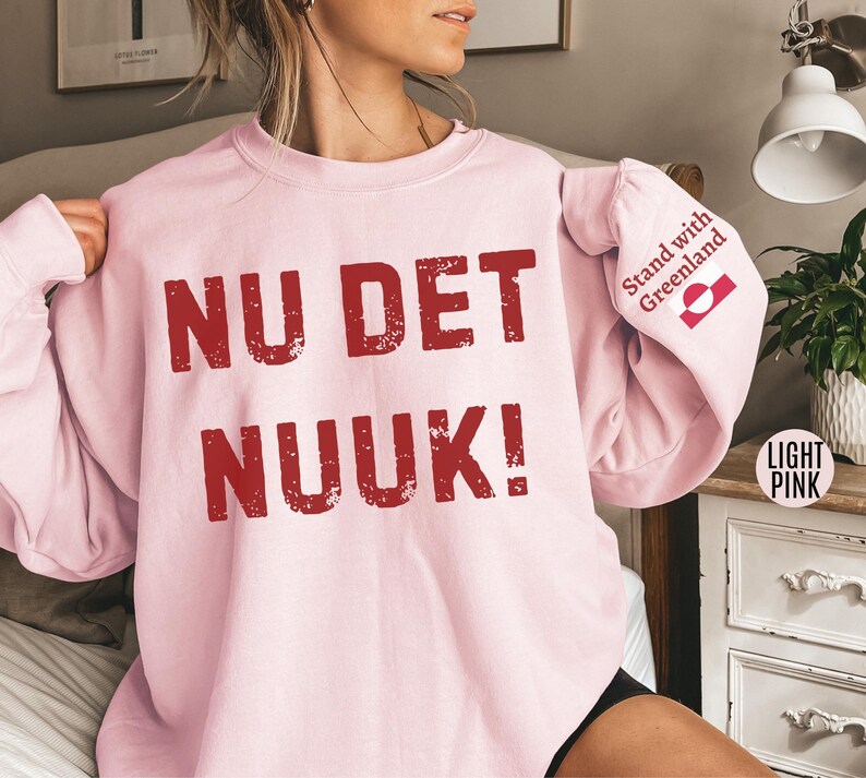 May include: Light pink sweatshirt with the words "NU DET NUUK!" in distressed red lettering across the front. The right sleeve has the text "Stand with Greenland" and a Greenland flag. The sweatshirt is a light pink color.
