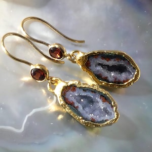 May include: A pair of gold-toned drop earrings featuring a small round dark red gemstone at the top and a larger, irregular geode slice at the bottom. The geode slice has a textured gold-toned edge, a dark brown outer ring, and a sparkling white center.