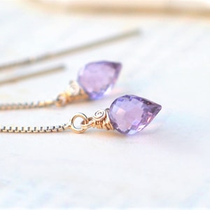 May include: Close-up of delicate earrings featuring teardrop-shaped, faceted lavender gemstones. The gemstones are suspended from fine, gold-colored chains. The earrings have a minimalist design, perfect for everyday wear or special occasions.