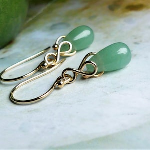 May include: A pair of drop earrings featuring smooth, light green teardrop-shaped stones suspended from gold-toned metal hooks. Each earring includes a delicate, looped wire design connecting the hook to the stone, creating a simple, elegant look.