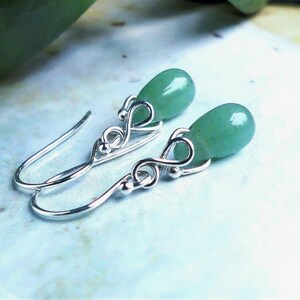 May include: A pair of drop earrings featuring smooth, teardrop-shaped green aventurine stones suspended from silver-toned metal hooks. The metalwork includes a delicate infinity-style loop design that connects the stone to the ear wire.