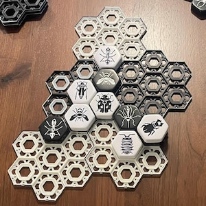 May include: A white and black board game with hexagonal tiles and pieces. The pieces are black and white and feature different insect designs. The game is set up on a wooden table.