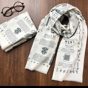 May include: Off-white scarf with black text and decorative elements, including the text "PSALM ONE TWENTY ONE". The scarf is draped and folded, with a fringed edge. A pair of round eyeglasses rests nearby.