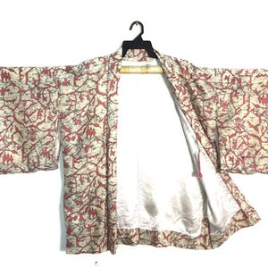 May include: A haori jacket featuring a cream-colored base with a repeating pattern of red and grey vine-like motifs. The garment has wide sleeves, an open front, and a plain white interior lining, displayed on a black plastic clothes hanger.