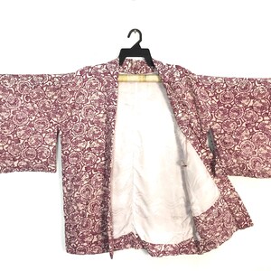 May include: A haori jacket featuring a repeating maroon and white circular floral pattern across the exterior. The garment has wide sleeves and an open front, revealing a plain white interior lining. It is displayed on a black plastic hanger.