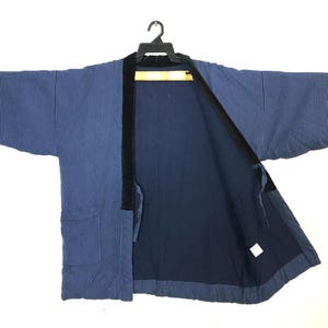 May include: A blue and white striped Japanese kimono jacket. The jacket has a dark blue interior and black trim along the collar and front edges. It features two front pockets and is hanging on a black hanger.