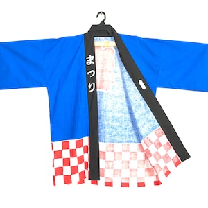 May include: A blue Japanese festival coat, or happi coat, with black trim and a red and white checkered pattern at the bottom. The coat has Japanese text on the front. The coat is hanging on a black hanger.