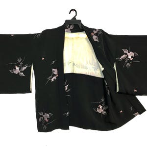 May include: A black kimono jacket featuring a delicate floral pattern with muted pink and grey leaves and blossoms. The garment has wide sleeves and a light-colored interior lining, displayed on a black plastic hanger against a plain background.