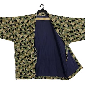 Puede incluir: Chaqueta estilo kimono con estampado de camuflaje y interior azul marino. La chaqueta presenta un diseño de camuflaje verde, marrón y negro en el exterior. Cuelga de una percha negra sobre un fondo blanco.