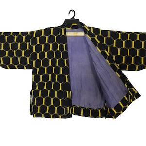 Peut inclure: Veste kimono à motifs noirs et jaunes avec un motif en nid d'abeille. La veste est ouverte, révélant une doublure violet clair. Les manches sont larges et le vêtement est suspendu à un cintre noir.