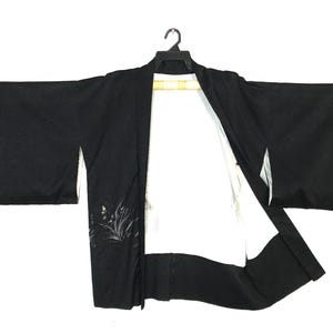 May include: A black haori jacket featuring wide sleeves and a white interior lining. The exterior fabric displays a subtle, dark-toned botanical pattern near the hem. The garment is displayed on a black plastic hanger against a plain white background.
