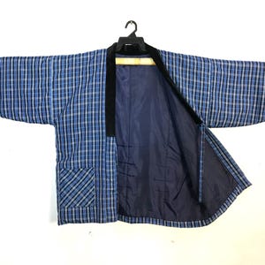 May include: A blue and white checked kimono jacket with black trim and a navy blue lining. The jacket has wide sleeves and a pocket. It is hanging on a black hanger.
