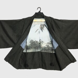 May include: A dark grey kimono, open to reveal a landscape design of a mountain and trees on the lining. The kimono is suspended from a black hanger. The interior design features a detailed illustration of a mountain and trees.